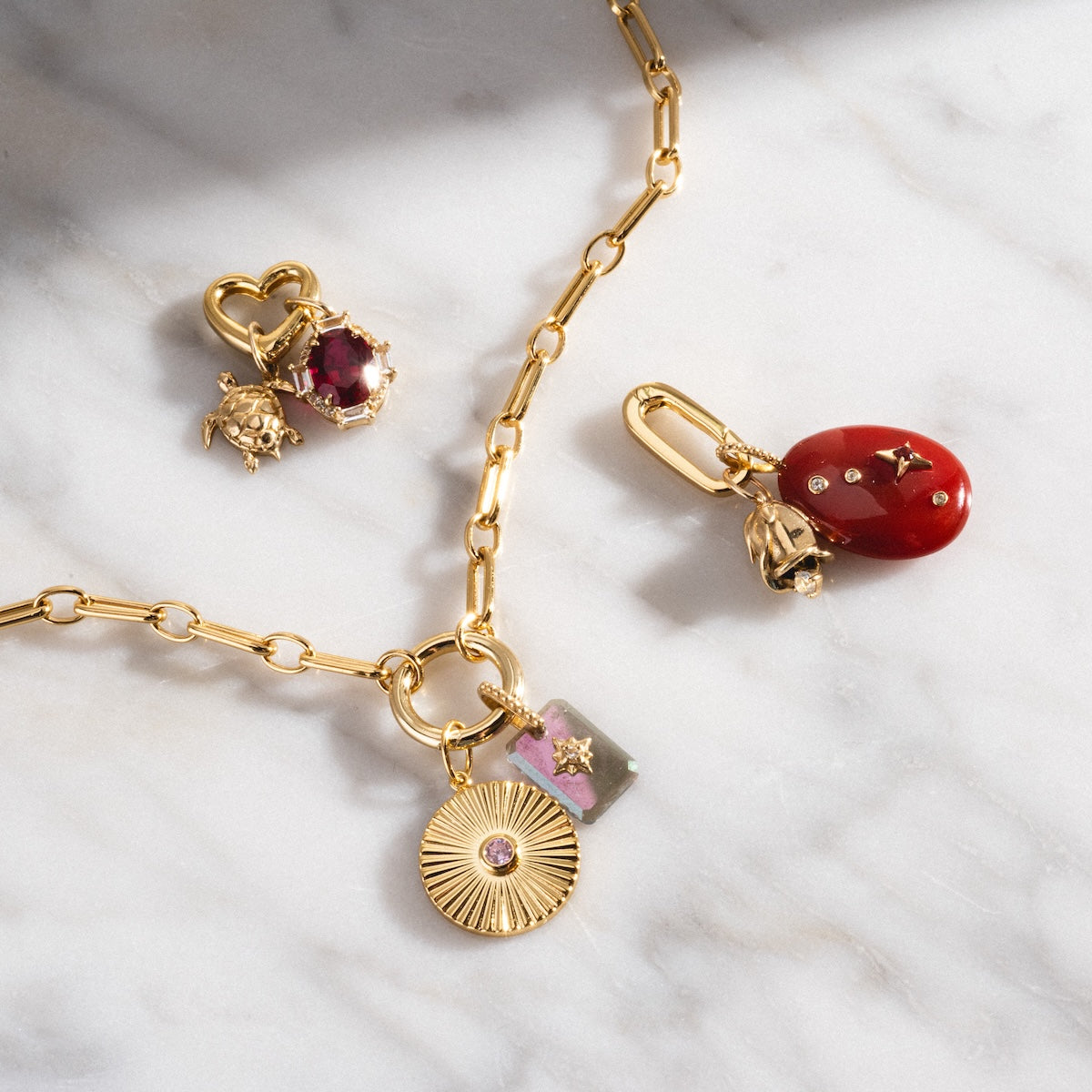 The Perfect Start Charm Holder Necklace, featuring a gold-plated chain with a sunburst disc, floral rectangle charm, and pink stone pendant, rests on white marble beside two gold and gemstone charms—an elegant, customizable accessory.