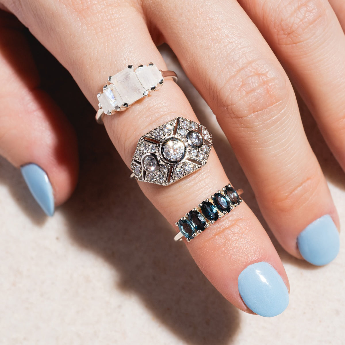 A hand with light blue nail polish wears three rings: a sterling silver ring with a clear rectangular stone on the index finger, the Venus Ring—an ornate vintage-inspired circular diamond ring—on the middle finger, and a dark gemstone band on the ring finger.