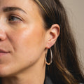 A close-up of a woman with long brown hair and clear skin, facing slightly left, highlights her ear adorned with a large, shiny teardrop-shaped Fluid Hoop Earring.