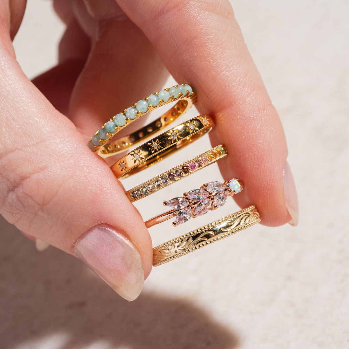 A manicured hand displays five gold rings, including the Solid Gold Filigree Ring, each adorned with gemstones, engraved details, and sparkling stones—all set against a light background.
