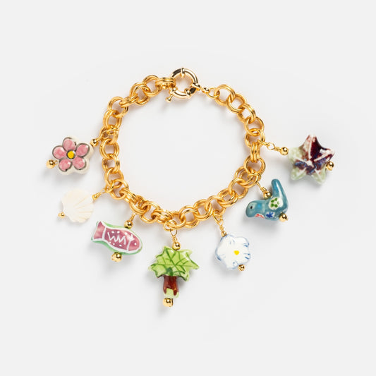 The Good Idea Bracelet is a 24k gold plated charm bracelet with seven colorful porcelain charms: pink flower, white shell, red fish, green tree, blue bird, blue and white shape, and purple star.