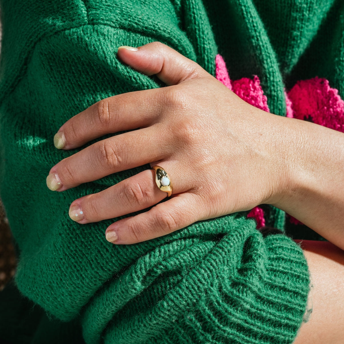 A person wearing a green sweater with pink letters crosses their arms, showing off the Aether Opal Ring Sample Size 6 (5 mm opal, recycled brass) on their finger. Their fingernails are short and natural.