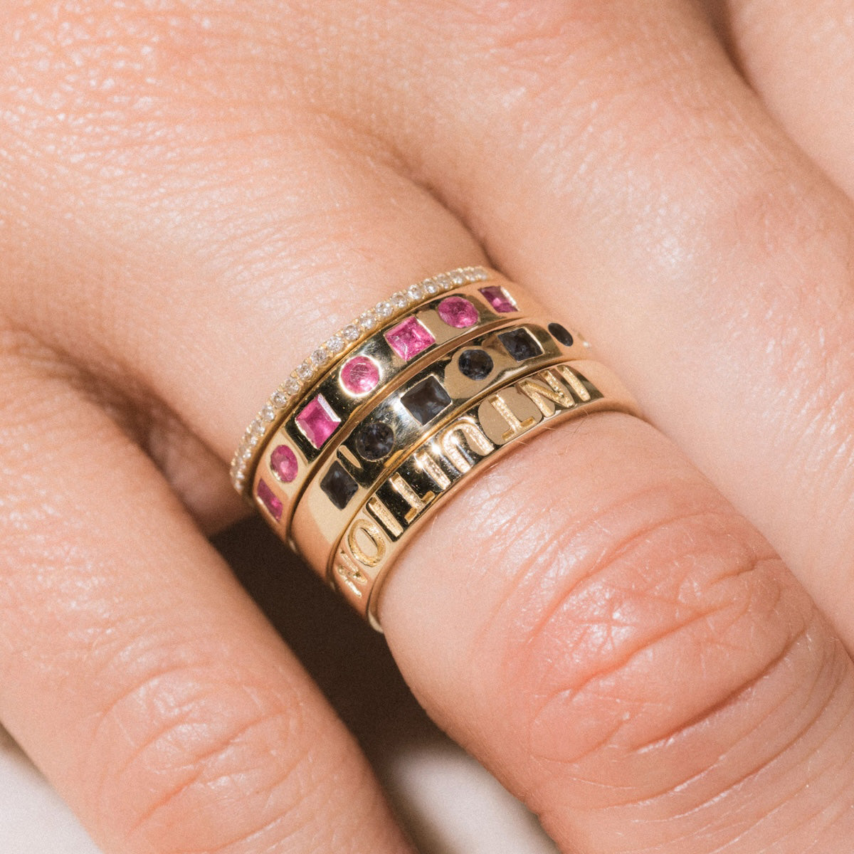 A close-up of a hand wearing four stacked Solid Gold Within You Birthstone Band rings in 9k yellow gold, size 7—some set with natural pink and black stones, and one 3.1 mm band engraved with NOT QUITE OURS.