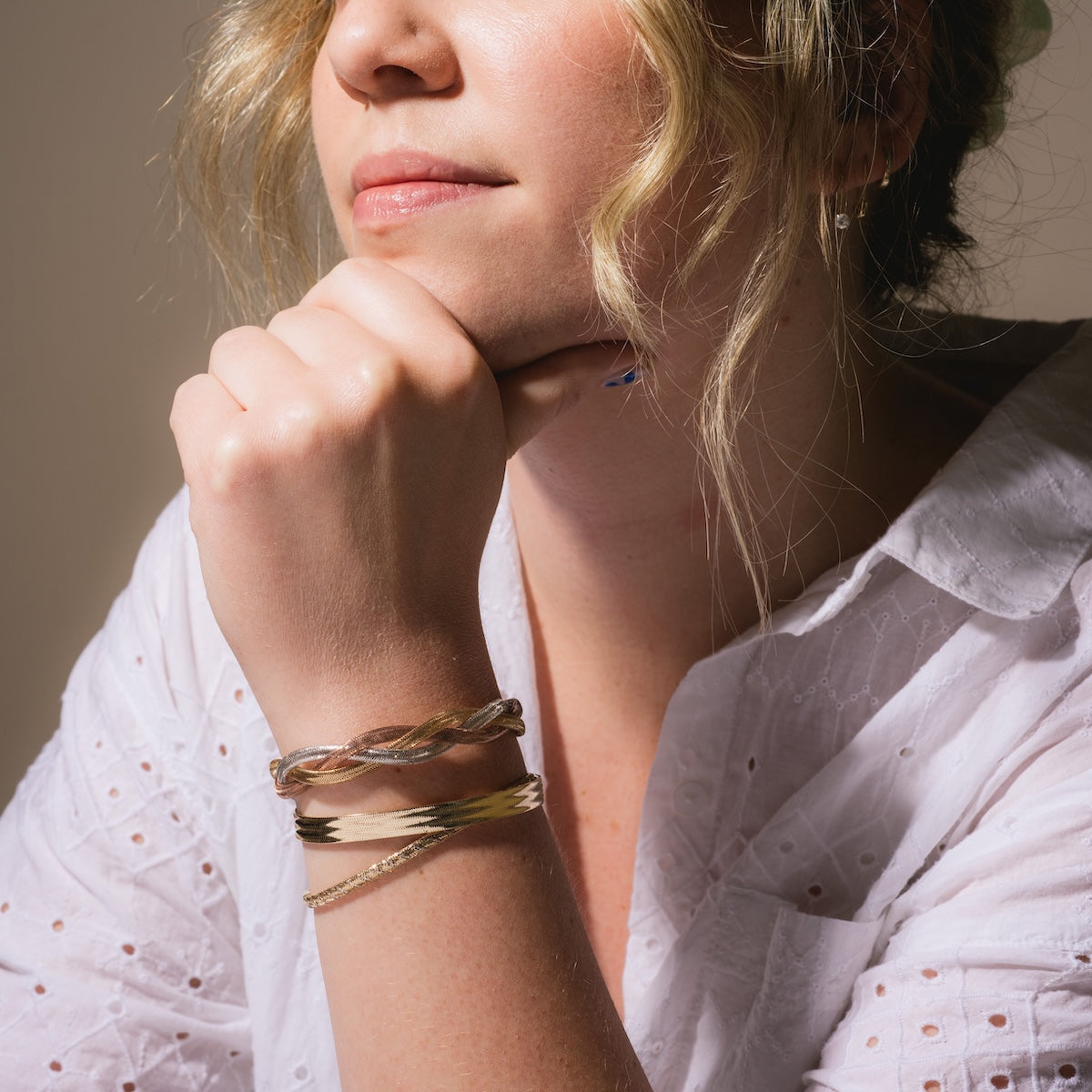 A woman with wavy blonde hair, dressed in a white textured blouse, rests her chin on her hand. On her wrist is the Solid Gold Mixed Metal Braided Mesh Bracelet Sample, highlighted by soft lighting that accents her relaxed and thoughtful pose.