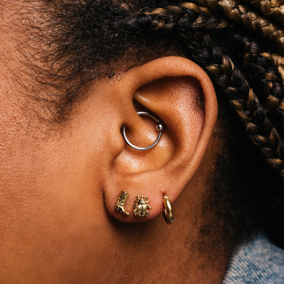A close-up of a person with braided hair wearing a blue shirt and multiple piercings, including charming Cowboy Boot Studs in the lobe, captures Local Eclectic style.