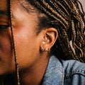 A close-up shows Lady Bug Studs and other 14k gold plated hoop earrings on medium brown skin, with braided hair featuring blonde highlights, paired with a blue denim jacket.