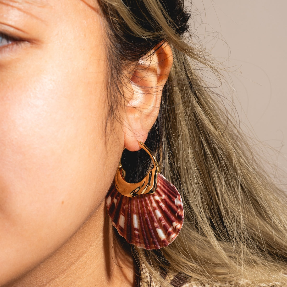 Close-up of a person with light brown skin and long, light brown hair wearing the Mira Earrings Sample—14k gold plated hoops featuring a maroon and white natural shell charm.