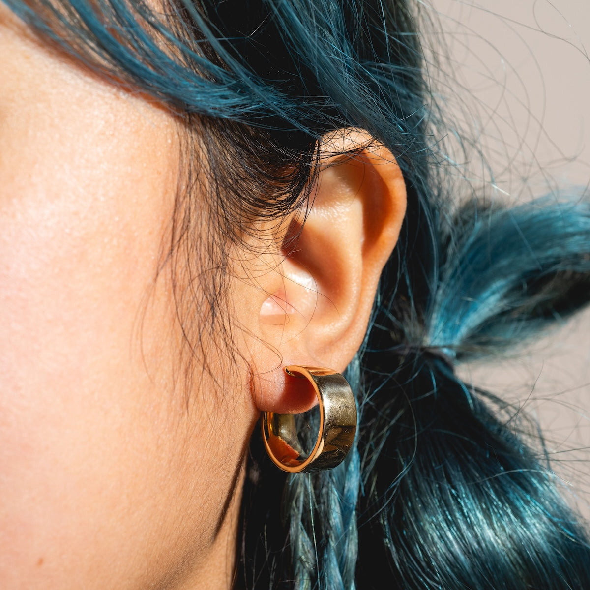 Close-up of a person’s ear with blue hair, wearing Solid Gold Flat Chunky Hollow Hoops in White Gold Sample. Hair is tied back, and the softly blurred background highlights the elegant 19.5 mm accessory.