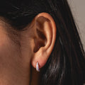 Close-up of a persons ear with straight dark hair, wearing the Opal Ombre Deux Huggies—small, shiny silver earrings. The softly blurred neutral background adds emphasis to the jewelry.