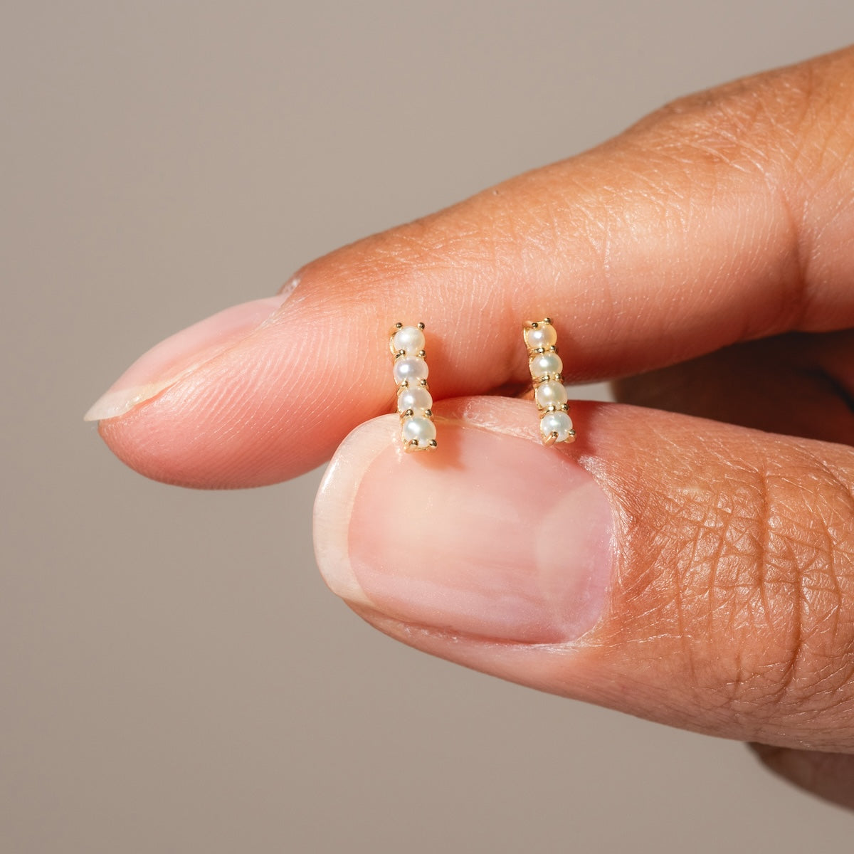 A hand holds Solid Gold Pearl Bar Flat Back Studs, each earring featuring a vertical row of four tiny white pearls. The neutral, blurred background keeps the focus on the elegant gold and pearl design.