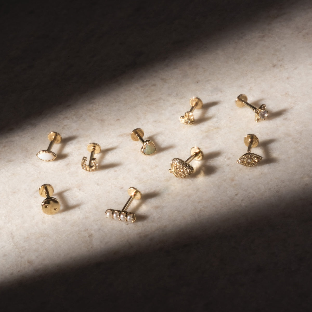 Nine Solid Gold Opal Marquise Flat Back Studs with various designs, featuring opal accents and textured patterns, are artfully arranged on a stone surface in soft, natural light.