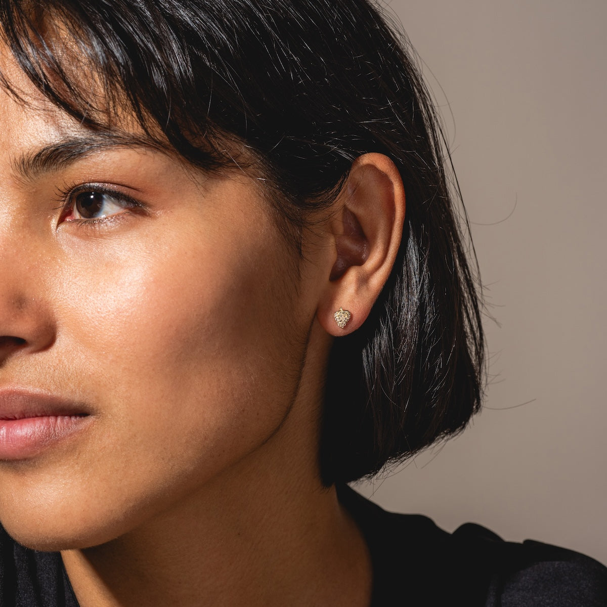 A close-up of a woman with short dark hair wearing Solid Gold Strawberry Flat Back Studs in her left ear. She has clear skin, a neutral expression, and faces slightly left against a plain background.