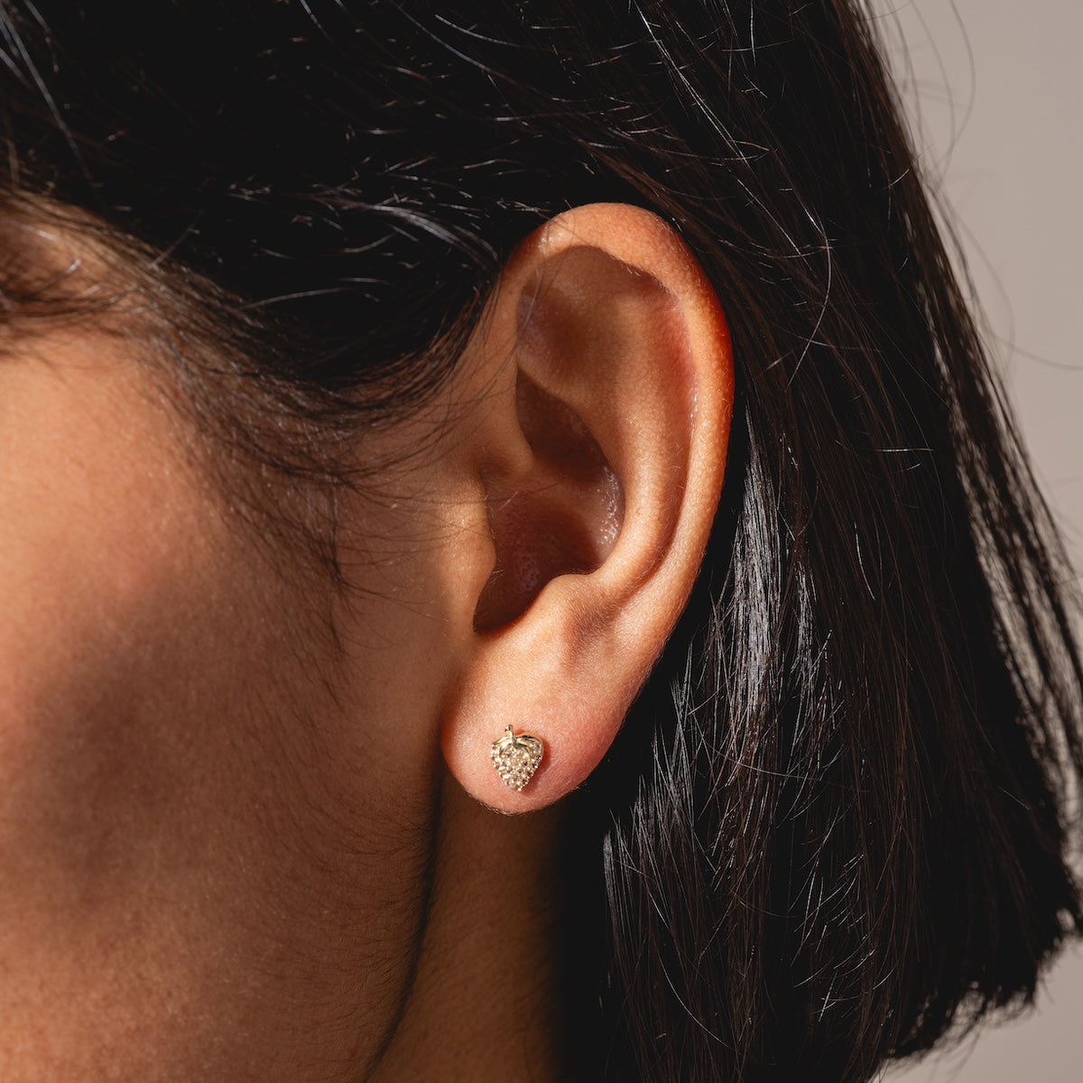 Close-up of a person’s ear with short dark hair, wearing a Solid Gold Strawberry Flat Back Stud earring, heart-shaped and adorned with tiny clear stones.