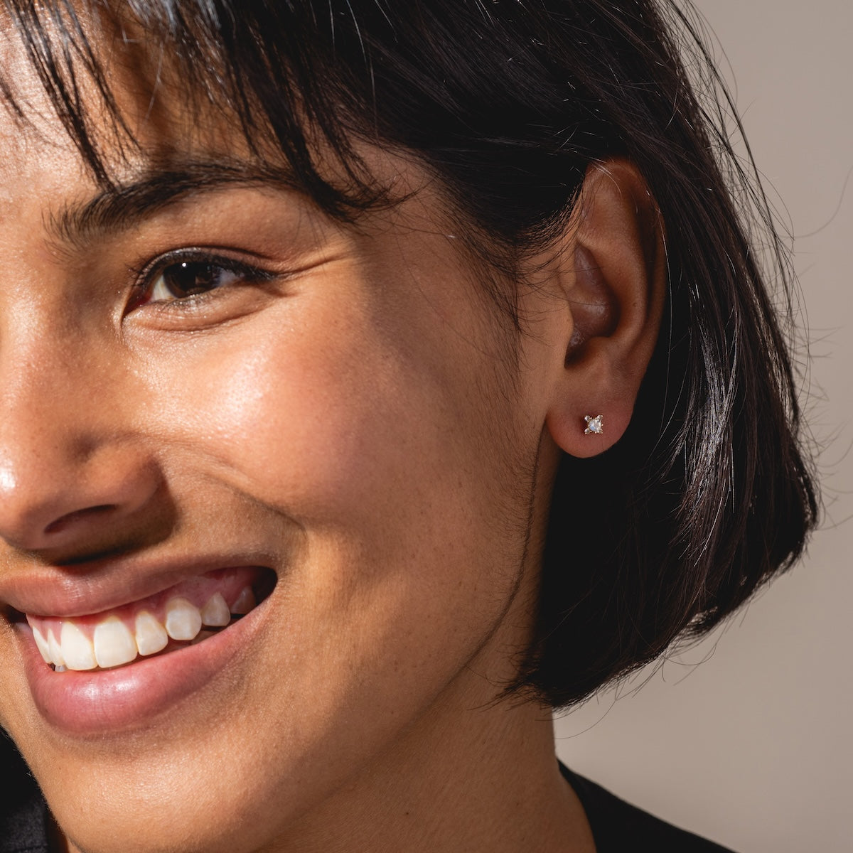 A woman with short dark hair smiles, revealing white teeth, while wearing a small Solid Gold Moonstone Flat Back Stud in her left ear. The bright image is focused on the left side of her face.