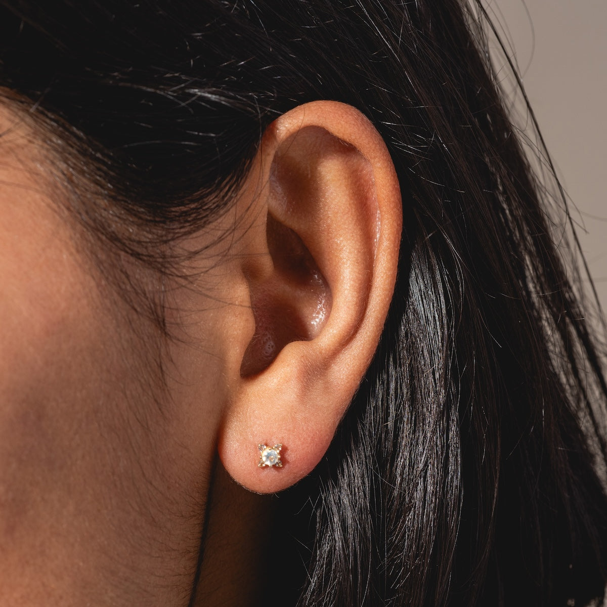 Close-up of a person’s ear with straight, dark hair, wearing a small Solid Gold Moonstone Flat Back Stud in the earlobe. The neutral, blurred background highlights the elegant design of the earring.