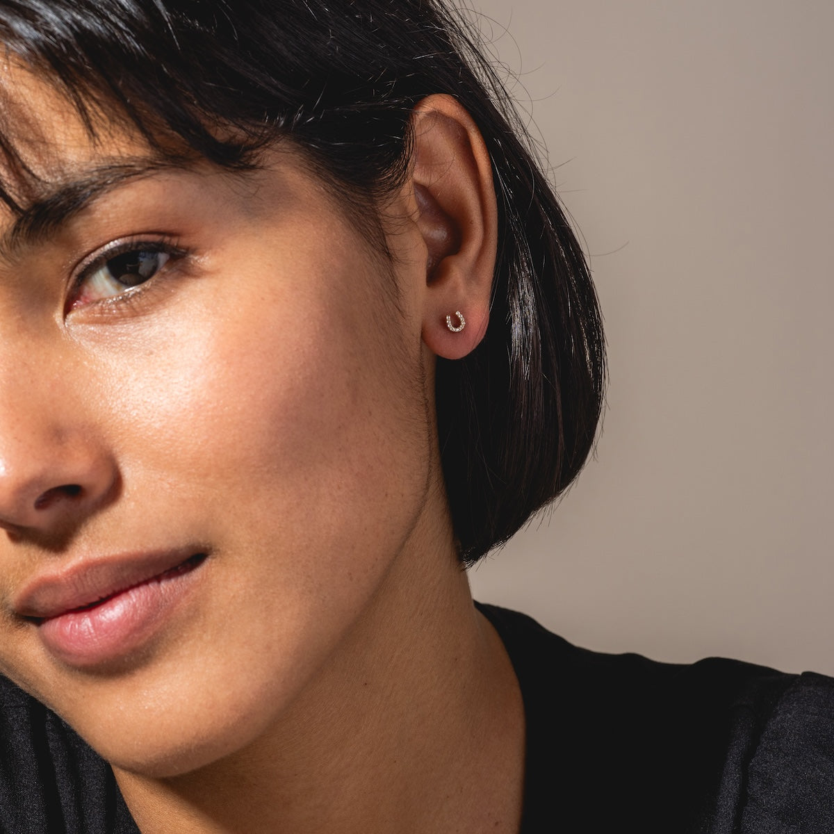 A close-up of a woman with short dark hair wearing Solid Gold Horseshoe Flat Back Studs in her left ear. She has smooth skin, is looking slightly past the camera, and is dressed in a black top.