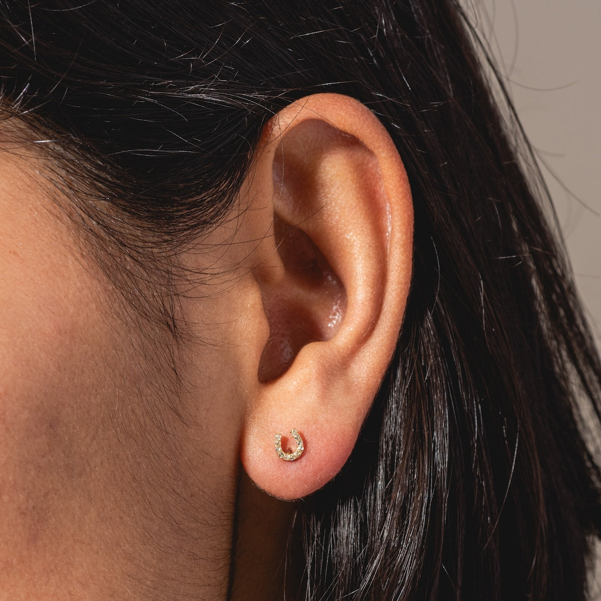 Close-up of a person's ear with straight dark hair, wearing Solid Gold Horseshoe Flat Back Studs featuring a single clear gemstone. The background is neutral and softly blurred.