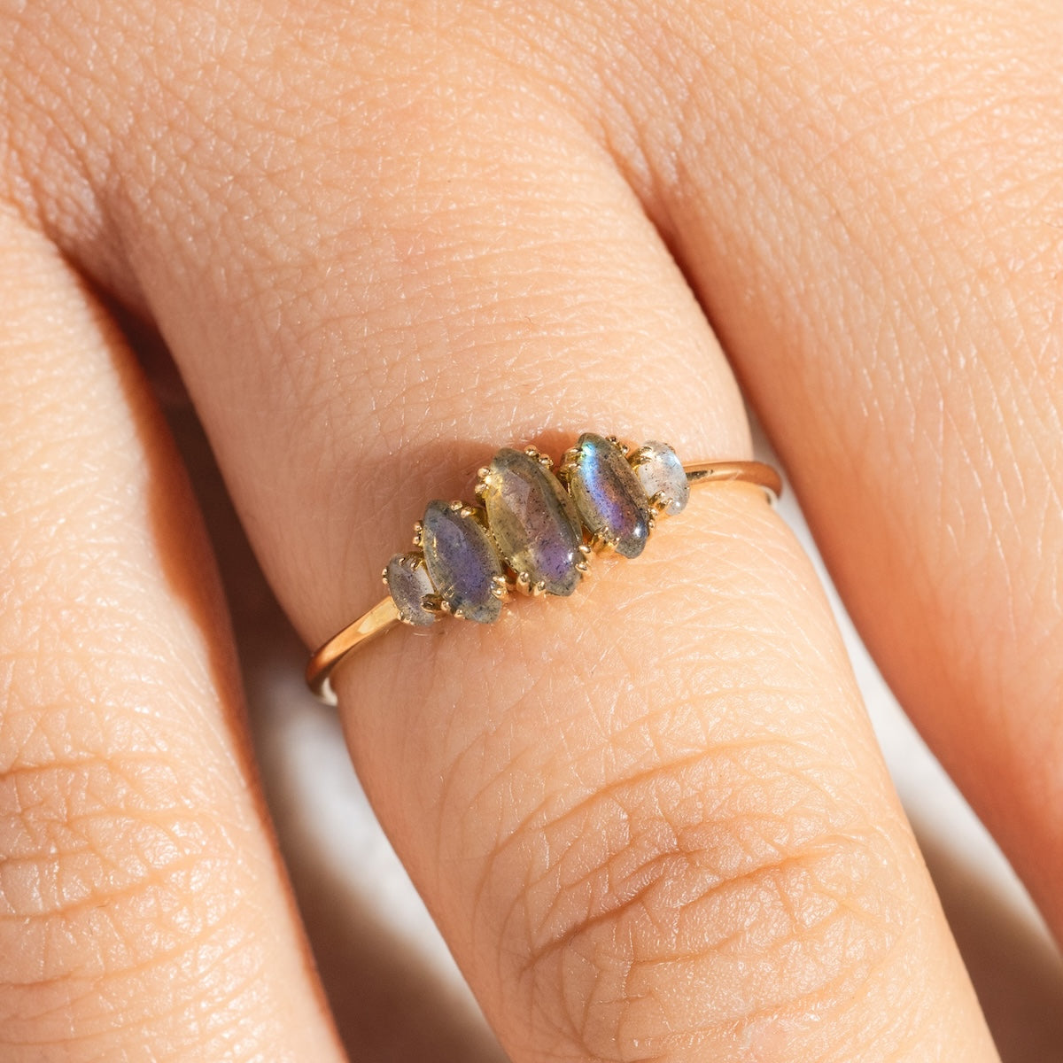 A close-up of a light-skinned hand wearing the Solid Gold Labradorite Marquise Cluster Ring, featuring five small, marquise-cut iridescent gemstones set in a row. Skin texture and lines are visible.