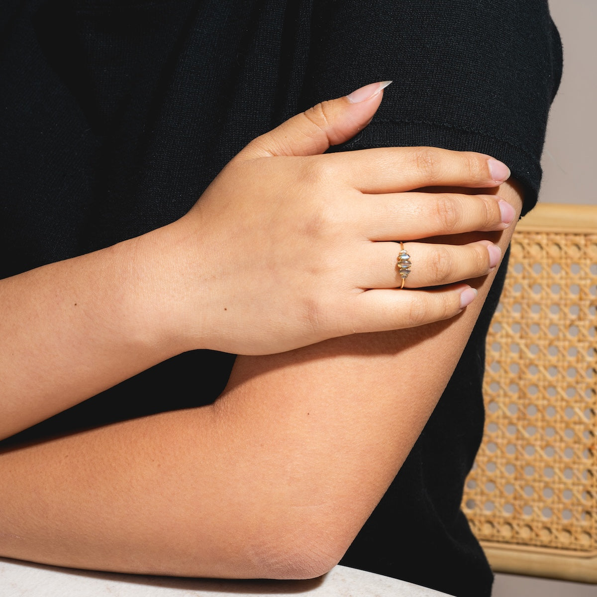 A person wearing a black short-sleeve top rests their left hand on their right arm, showcasing the Solid Gold Labradorite Marquise Cluster Ring on their finger. The background features a woven chair and a white surface.