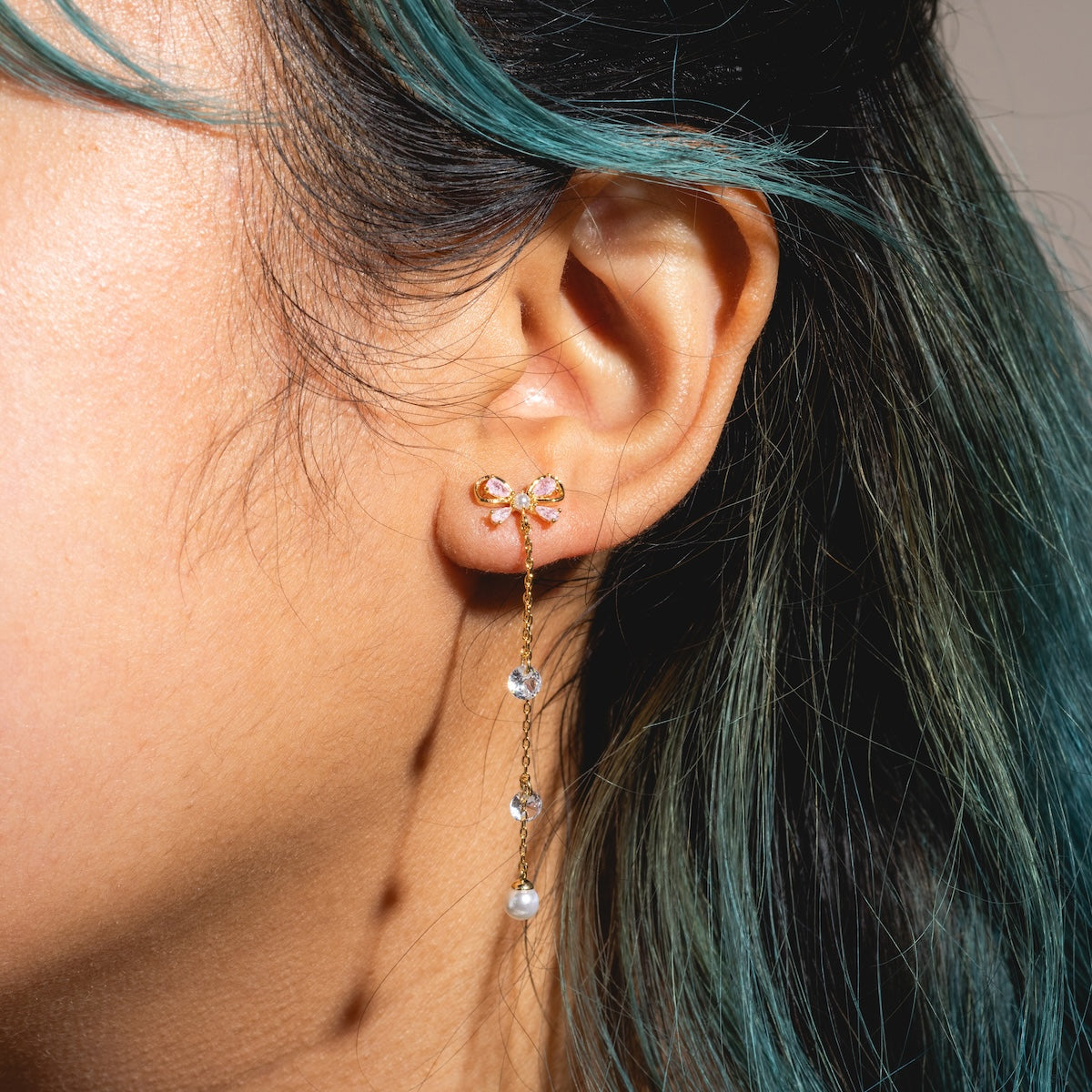 Close-up of a womans ear with teal-streaked hair, wearing Princess Pearl Earrings Samples—18k yellow gold plated, featuring a bow design with sparkling cubic zirconia crystals and a pearl at the end.
