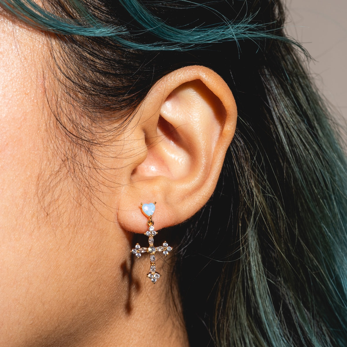 Close-up of a person’s ear wearing Serendipity Cross Dangle Earrings Sample—blue stone and jeweled cross pendant, crafted from 18k yellow gold plated brass with sparkling CZ accents. Teal-highlighted hair frames the ear.