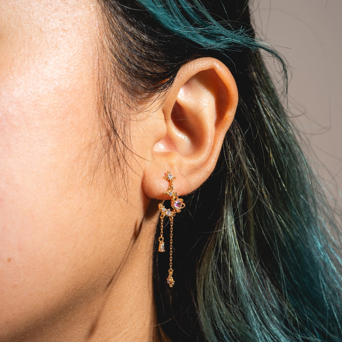 Close-up of a person’s ear with teal hair, wearing the Moonbeam Dangle Earrings Sample—18k yellow gold plated, featuring small star and planet charms on delicate chains and sterling silver posts.