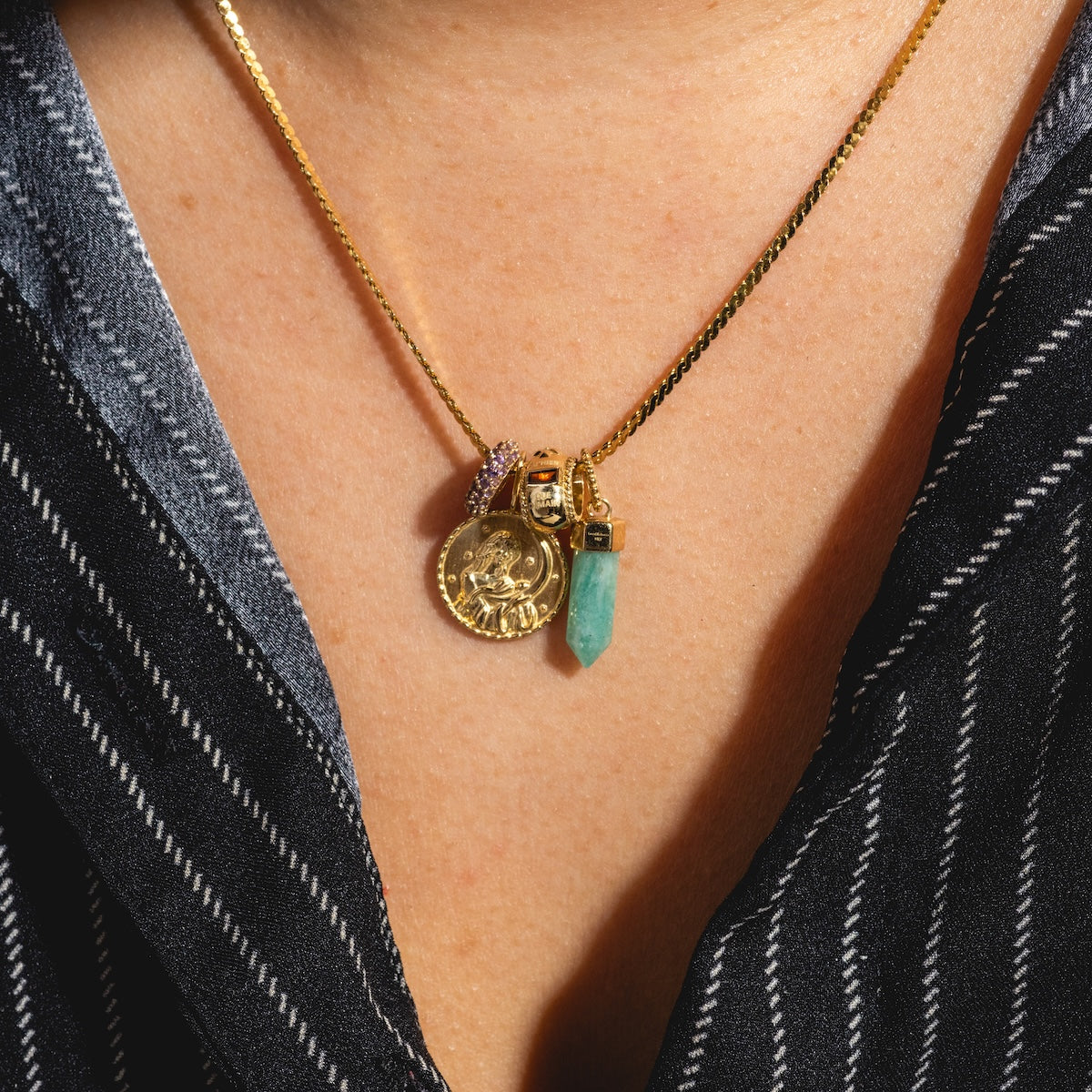 A close-up of a person in a black and white striped shirt wearing a 9k yellow gold chain with several pendants: a gold coin, turquoise crystal, the Solid Gold Qu'hier Que Demain Spacer Charm, and a jeweled ring.