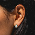 Close-up of a person with dark hair wearing Luster Studs—teardrop-shaped opal earrings set in 14k gold-plated metal and accented with small diamonds.