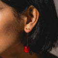 Close-up of a person with short dark hair wearing Cherry and Stem Earrings—yellow gold plated bar studs with long wires, featuring a red cherry dangling below the ear.