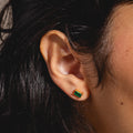 Close-up of a persons ear with straight dark hair, wearing Birthstone Baguette Studs—yellow gold vermeil earrings featuring a rectangular green gemstone. The image highlights these elegant studs against a neutral background.