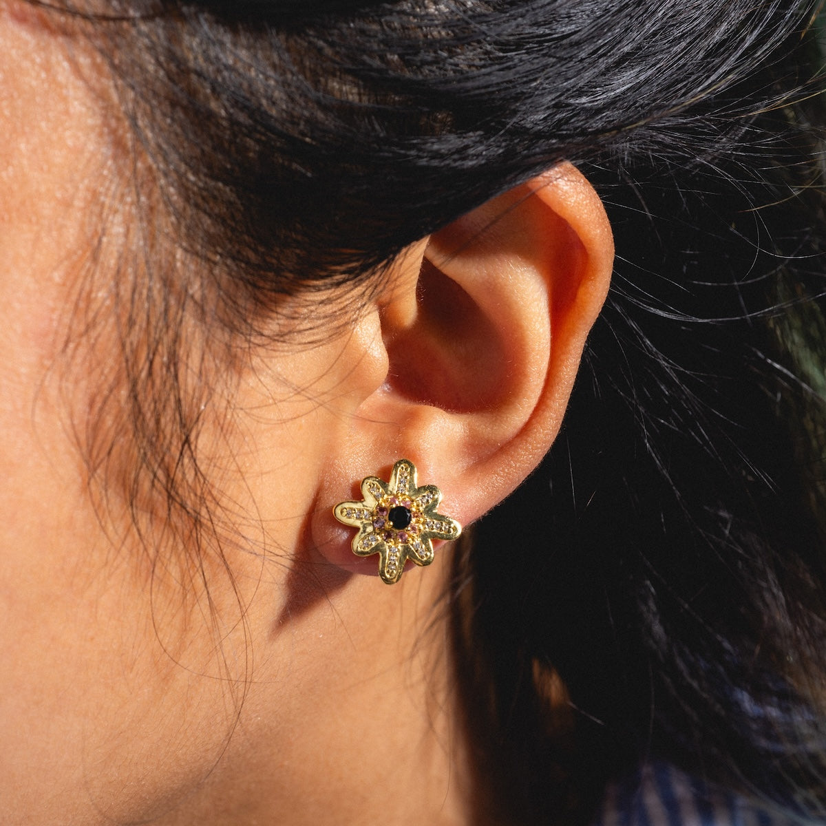 Close-up of a person wearing Wildbloom Stud Earrings Sample: gold flower-shaped studs with a black chalcedony center. Their dark hair is pulled back, and sunlight highlights the 18k yellow gold plating on the earring and ear.