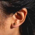 Close-up of a person’s ear wearing Boy Bye Earrings—small, gold-plated crown-shaped studs with tiny cubic zirconia gems. The person has dark hair and warm-toned skin.