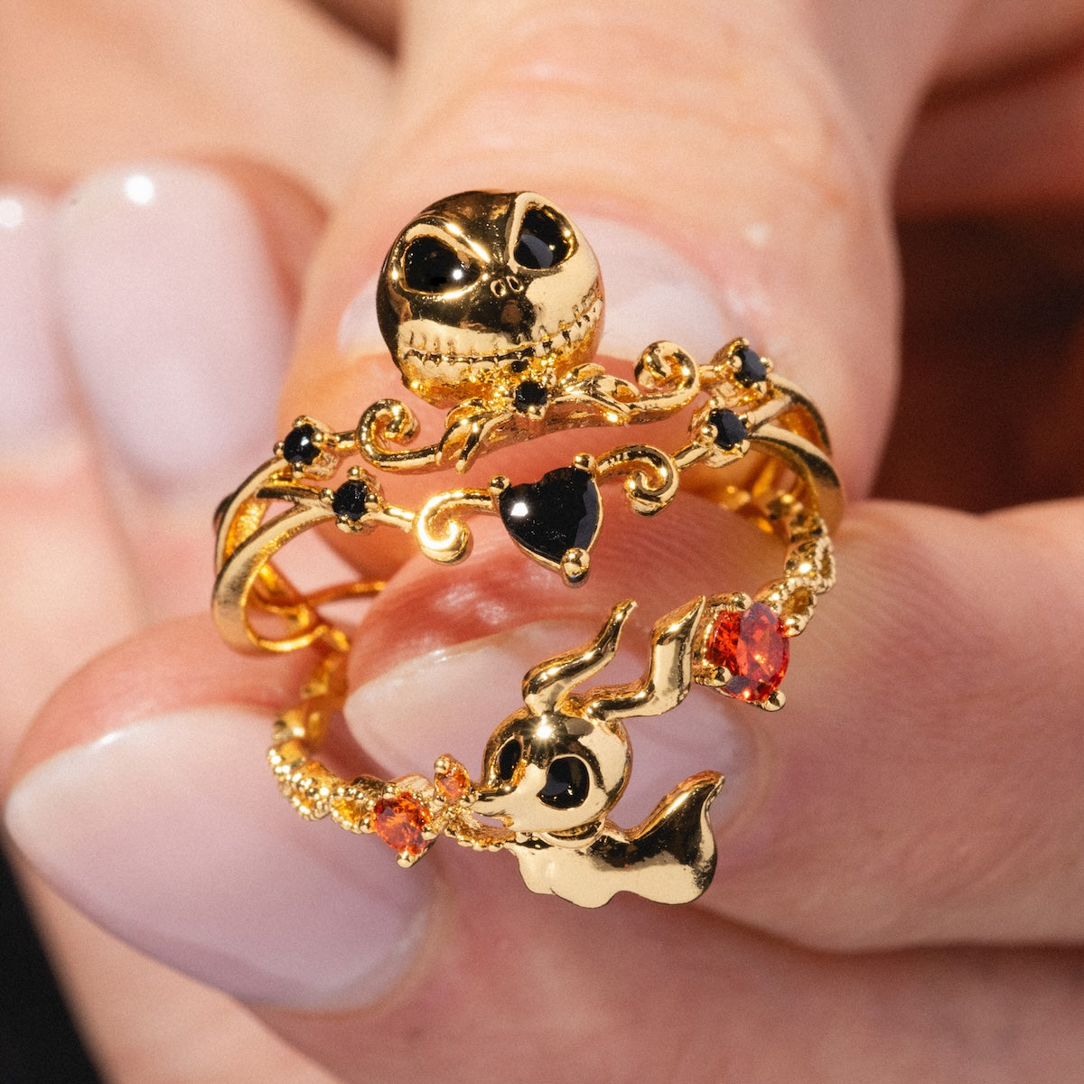 A hand with manicured nails holds two gold rings: a Nightmare Before Christmas ring with a skull and heart, and the Disney Zero Adjustable Ring shaped like a ghost dog, accented with orange and black gems—perfect for Disney fans.