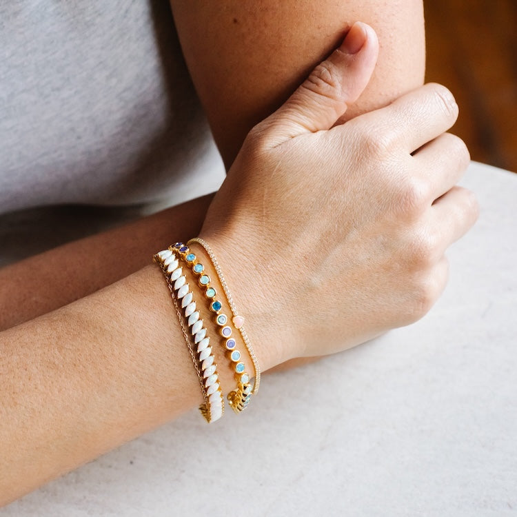 A person with light skin wears the Coral Heart Diamond Tennis Bracelet along with two other bracelets—a gold chain and a multicolored beaded one—while resting their arm on a white surface.