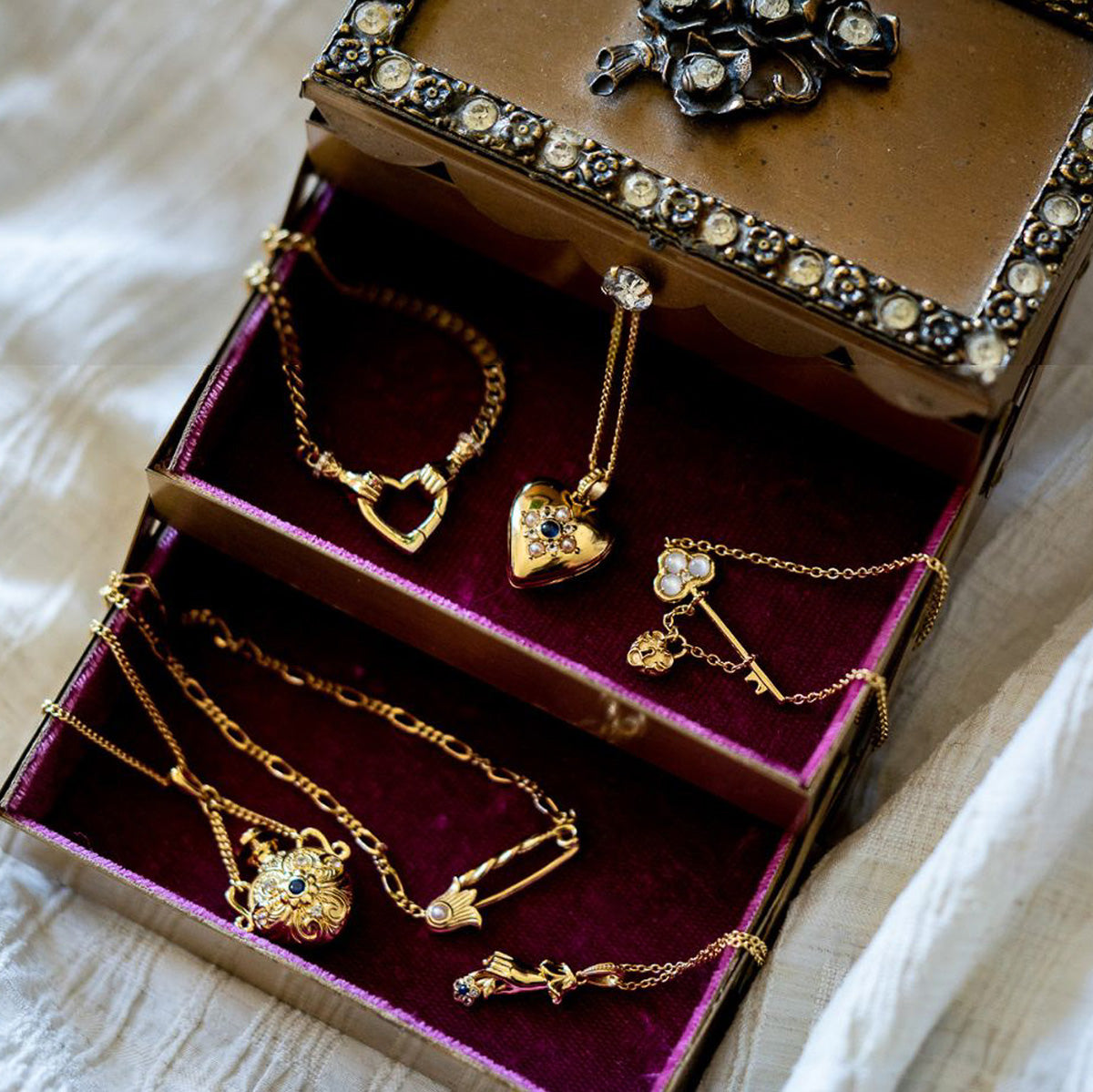 An ornate jewelry box with red velvet lining holds vintage gold necklaces and bracelets, such as the Forget Me Not Blue Topaz Memento Necklace and other heart-shaped and floral pendant pieces, displayed on a light fabric background.