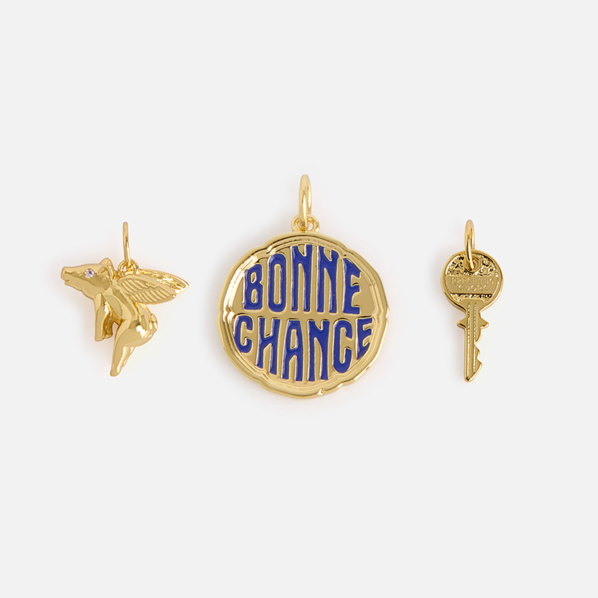 Charms by Mod + Jo features three gold-plated charms: a flying pig with wings, a round enamel “BONNE CHANCE” charm, and a tiny key-shaped good luck charm, all set against a plain white background.