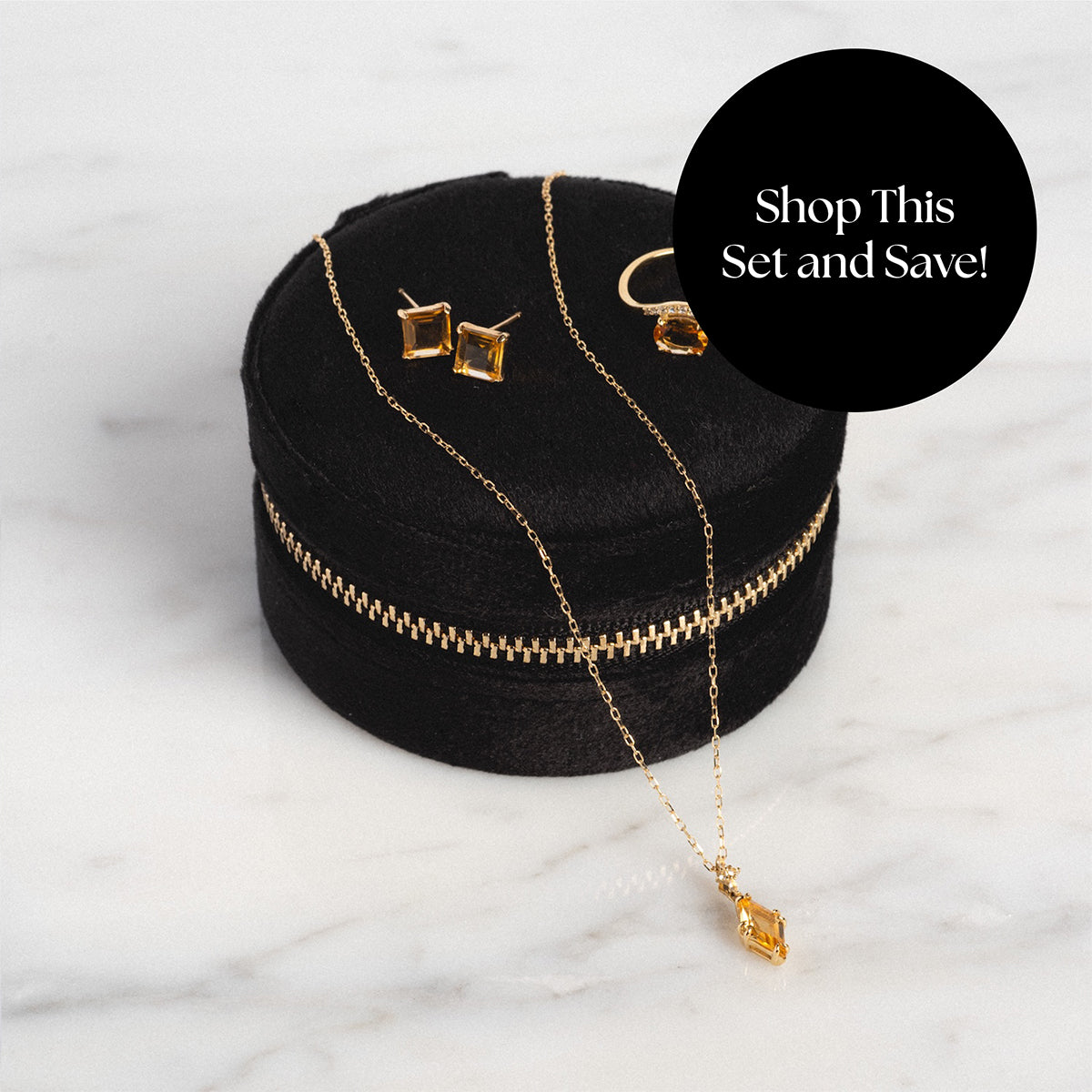 The Solid Gold 2025 November Capsule Citrine Statement Set—ring, necklace, and earrings with rectangular citrines—comes in a round black velvet case. “Shop This Set and Save!” is shown on a black circle with a white marble background. Includes free gift.