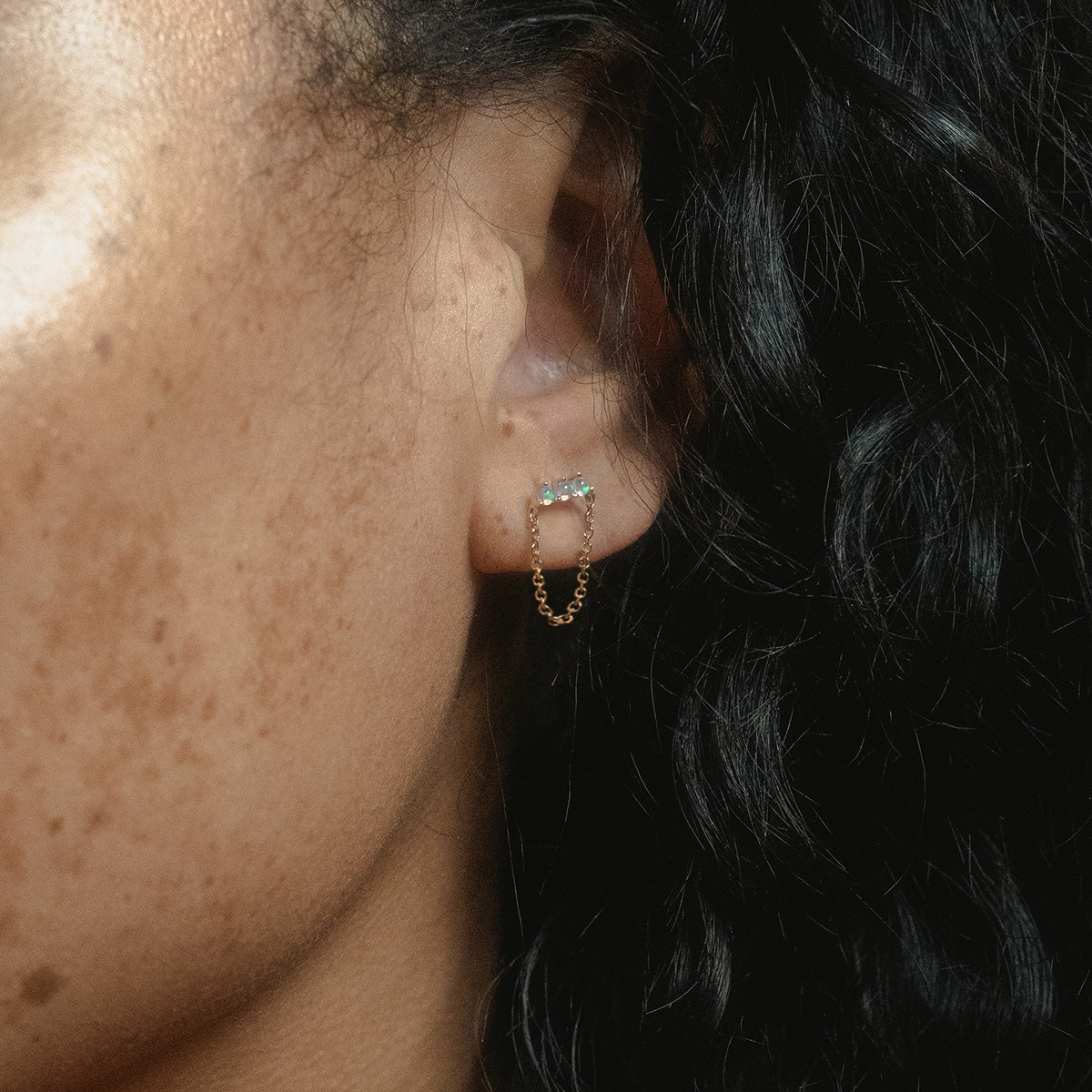 Close-up of a person’s ear wearing Solid Gold Opal Trio Chain Dangle Studs, with a trio of opals at the top and a delicate chain looping down. The person has curly dark hair and visible freckles.