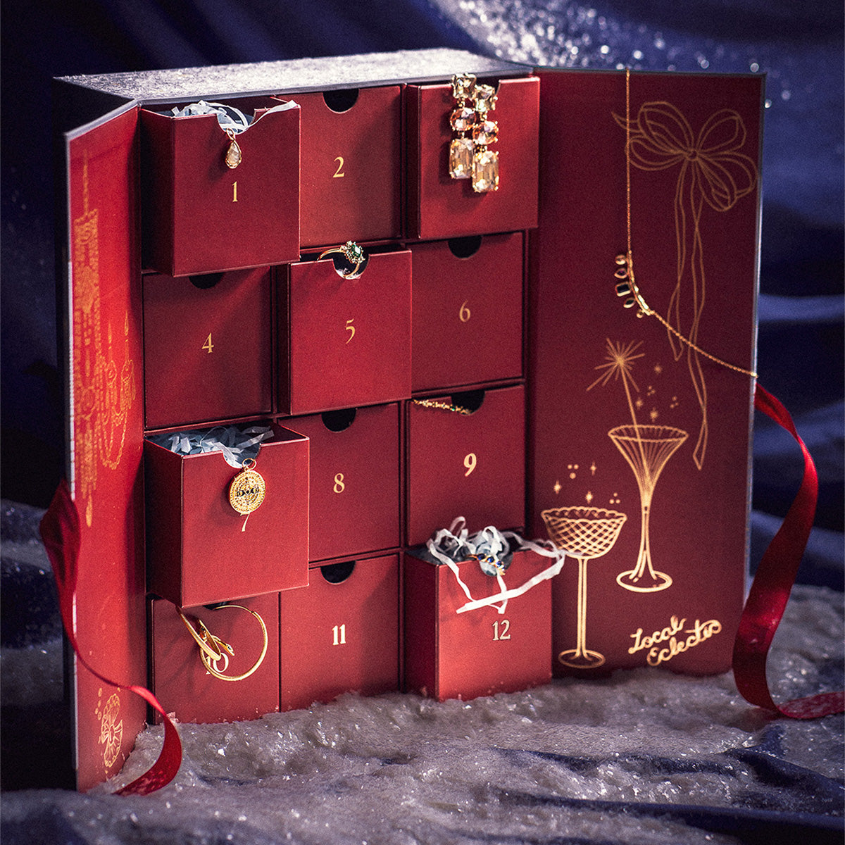 The 2025 Jewelry Advent Calendar, featuring twelve numbered drawers with assorted necklaces and earrings, sits open on snowy fabric, its red cover decorated with golden glasses and ribbon illustrations.