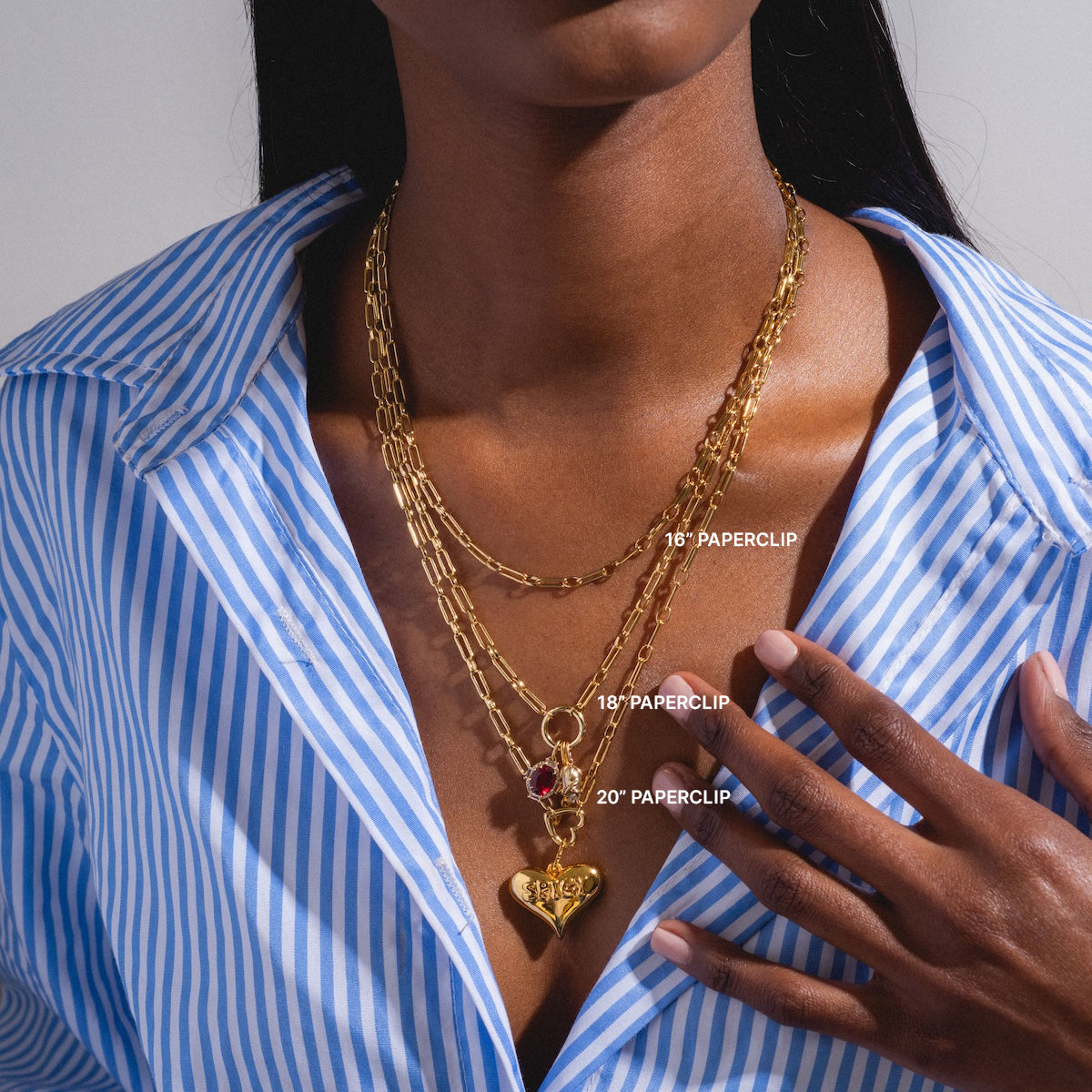 A woman in a blue and white striped shirt showcases the Perfect Start Charm Holder Necklace: three layered gold plated paperclip chains (16, 18, 20) with a heart-shaped pendant and red charm.