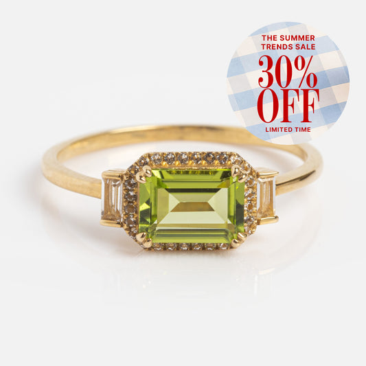 The Solid Gold Vintage Inspired East West Peridot Ring features a rectangular green gemstone, small clear stones, and side baguettes. Circular label: The Summer Trends Sale 30% OFF Limited Time. Family Gold elegance for every occasion.