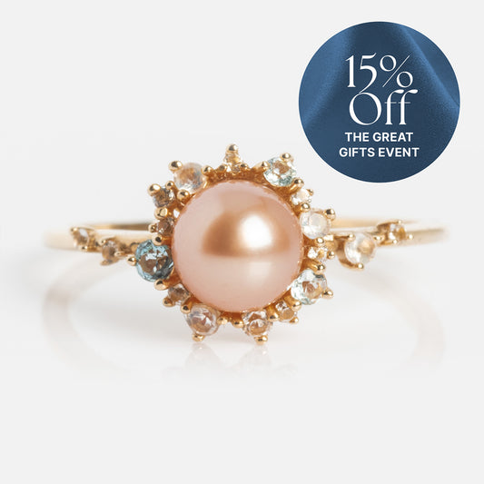 The 14k Pastel Pink Pearl Statement Ring showcases a central peachy-pink pearl, accented by clear and blue gemstones. A circular label in the top right reads, “15% Off THE GREAT GIFTS EVENT.”.