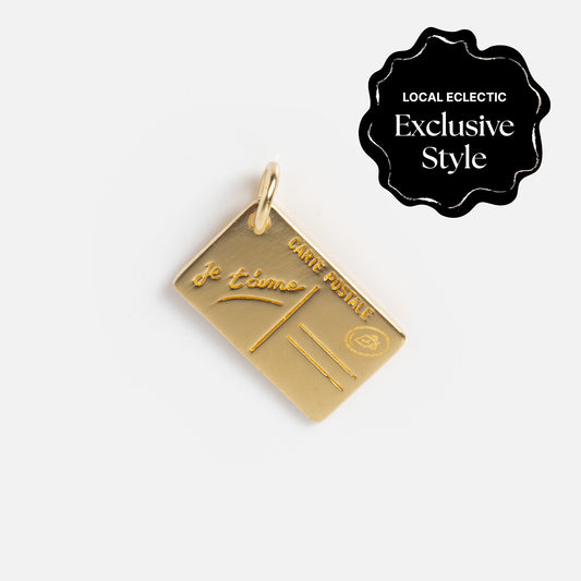 Gold postcard-shaped charm engraved with Je t’aime and CARTE POSTALE, inspired by travel. Features a black LOCAL ECLECTIC Exclusive Style badge. From the Zahava x Local Eclectic Globetrotter Charms collection.
