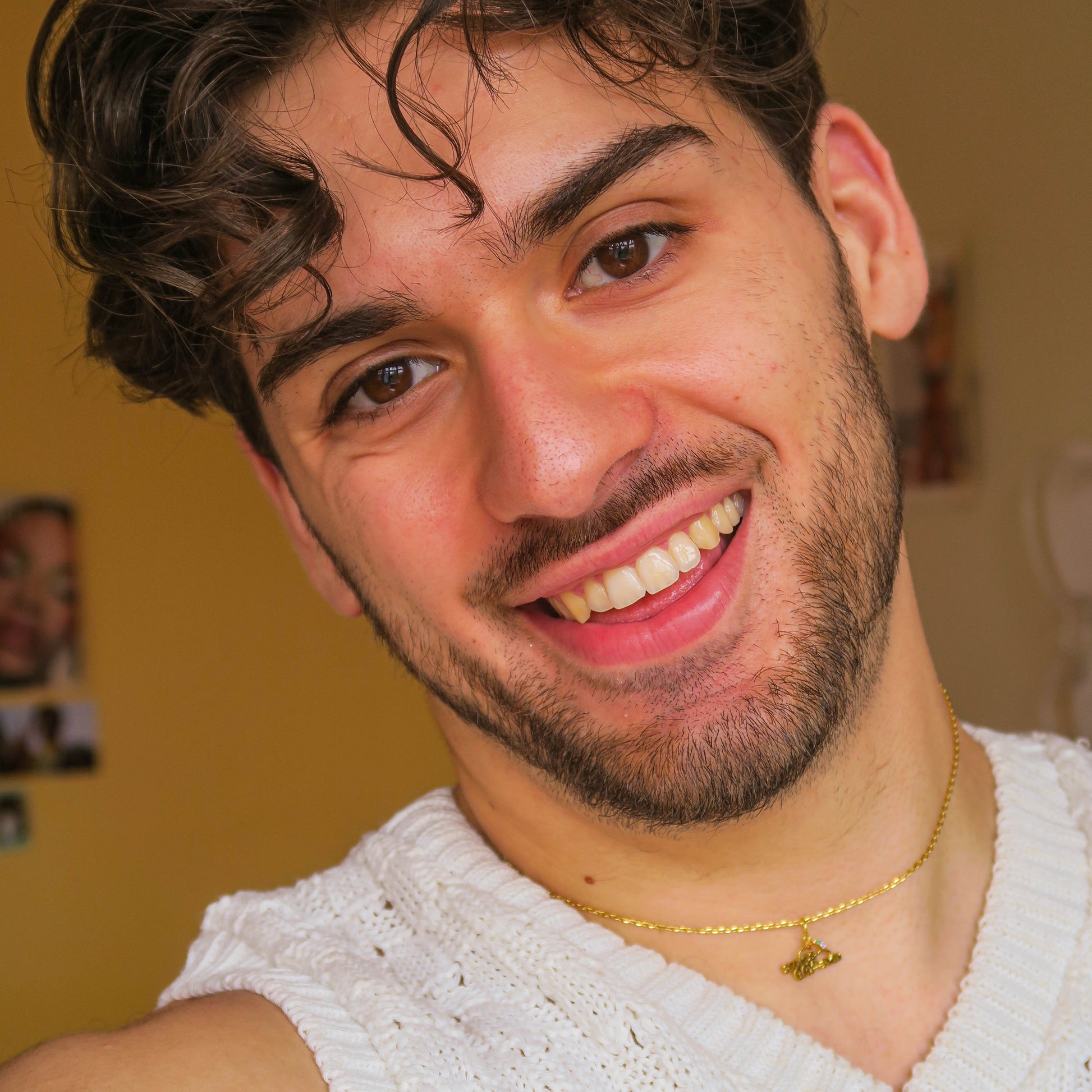 A young man with short curly hair and a beard smiles at the camera, wearing a white knitted top and a gold necklace featuring the Limited Edition PRIDE Month Charm, set against a softly lit yellow background with blurred photos.