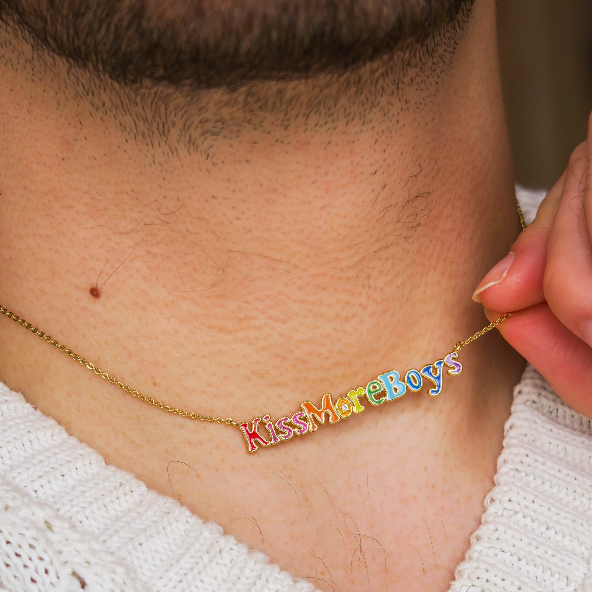 A close-up of a person's neck shows the Kiss More Boys Necklace, a gold chain with colorful letter charms spelling "Kiss More Boys." The person, wearing a white textured sweater, is holding the pendant.