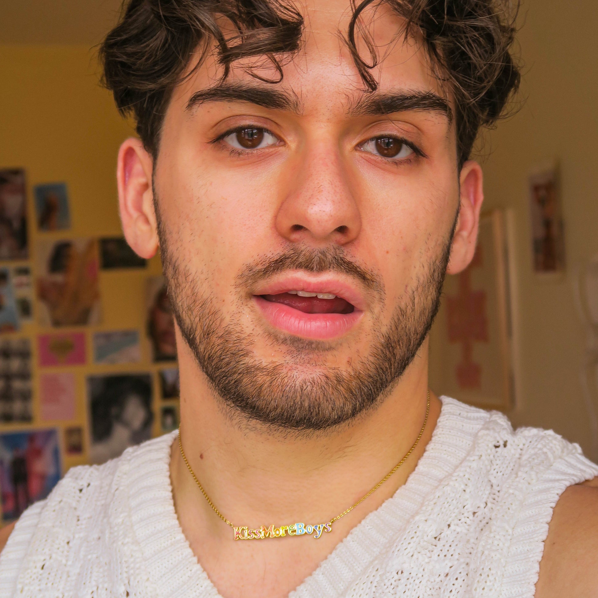A young man with wavy dark hair and a beard wears a white knit tank top and the Kiss More Boys Necklace, posing indoors against a yellow wall decorated with photos and posters.
