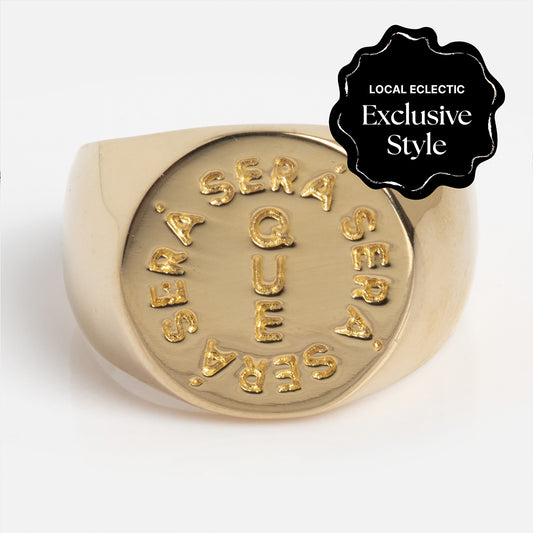 The I Like It Here Club x Local Eclectic Que Sera Sera Pinky Ring is a gold signet engraved with “QUE SERA SERA” in a circle, featuring a black badge in the corner marked “LOCAL ECLECTIC Exclusive Style.”.