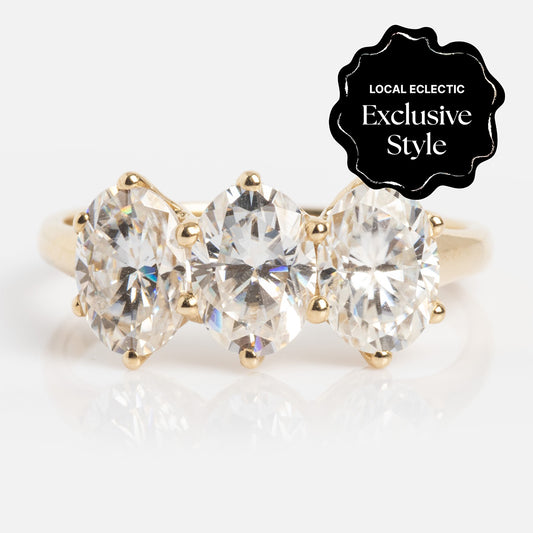 The Queenie Ring is a gold band with three large oval-cut clear Moissanite gems totaling 4.62 ctw, marked by a black "Local Eclectic Exclusive Style" label in the top right corner.