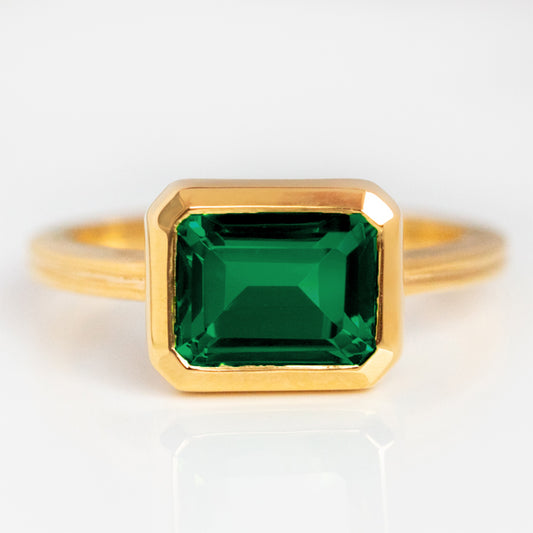 The East West Bold Bezel Ring Green Size 8 Sample features a rectangular, emerald-cut green gemstone in a simple bezel setting, shown on a white background.