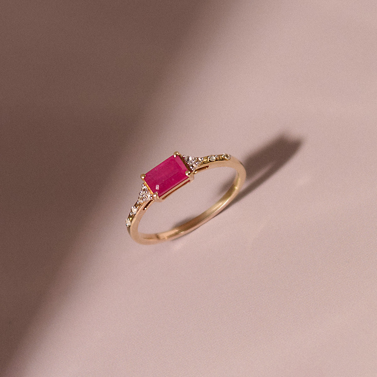 The Solid Gold Glimmering Birthstone Baguette Ring showcases a rectangular pink center stone with clear side accents, displayed on a soft pink surface—an elegant piece from Family Gold.